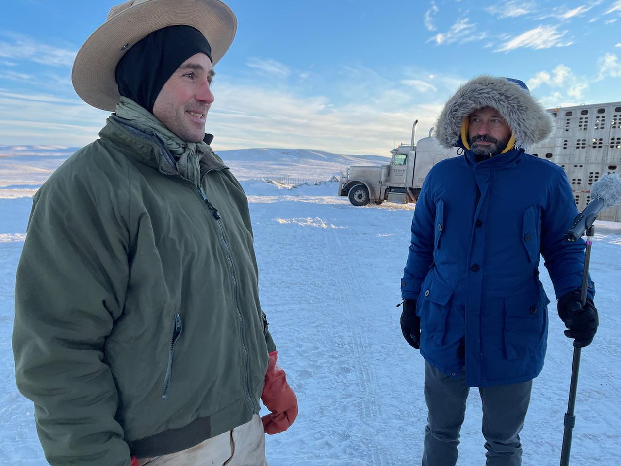Zwei Männer mit dicken Anoraks, Mützen und Handschuhen stehen unter blauem Himmel in einer Schneelandschaft.
