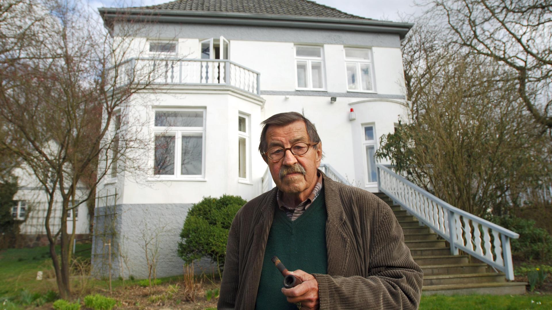 Günter Grass steht im Garten vor seinem Haus in Behlendorf. Grass hält eine Pfeife in der Hand. Das Haus ist zweistöckig. Eine Treppe führt aus dem Garten zum Haus. Günter Grass steht im Garten vor seinem Haus in Behlendorf. Grass hält eine Pfeife in der Hand. Das Haus ist zweistöckig. Eine Treppe führt aus dem Garten zum Haus.