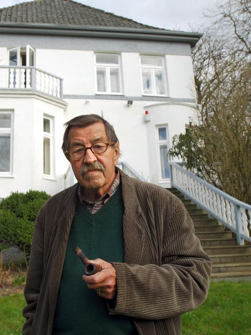 Günter Grass steht im Garten vor seinem Haus in Behlendorf. Grass hält eine Pfeife in der Hand. Das Haus ist zweistöckig. Eine Treppe führt aus dem Garten zum Haus. 