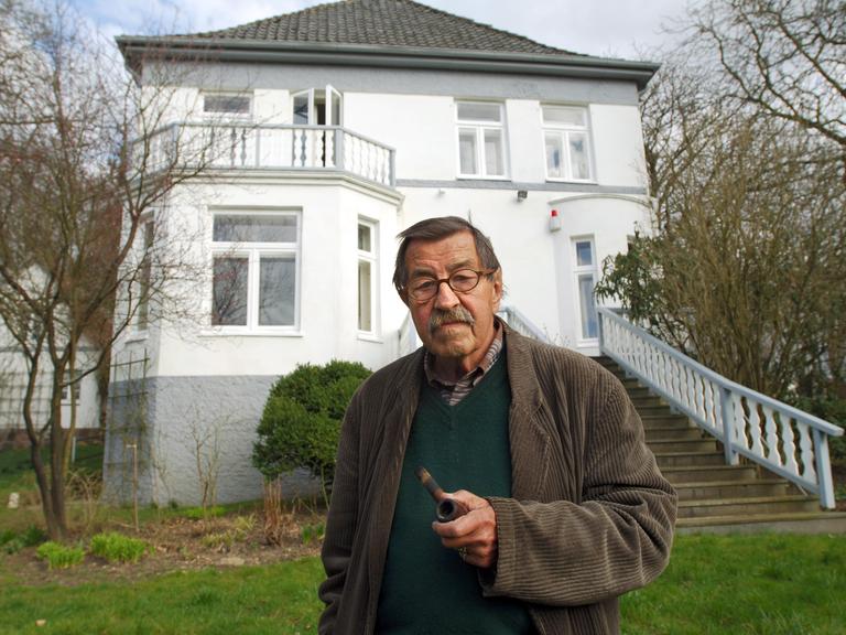 Günter Grass steht im Garten vor seinem Haus in Behlendorf. Grass hält eine Pfeife in der Hand. Das Haus ist zweistöckig. Eine Treppe führt aus dem Garten zum Haus. 