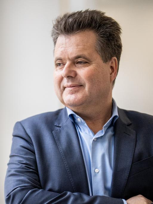 Ein Mann mit kurzen Haaren steht auf einem Flur an einem Fenster und schaut raus.