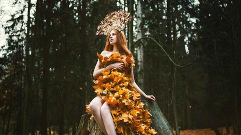 Eine Frau sitzt im Wald auf einem Baumstumpf und ist mit goldfarbenem Laub bedeckt, das aussieht wie ein Kleid.