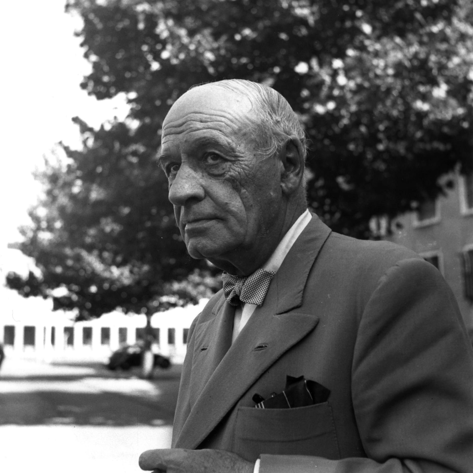 José Ortega y Gasset trägt ein graues Sakko und schaut zur Seite. Er steht vor einem Gebäude, im Hintergrund ist ein Baum und ein Auto zu sehen.