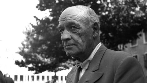 José Ortega y Gasset trägt ein graues Sakko und schaut zur Seite. Er steht vor einem Gebäude, im Hintergrund ist ein Baum und ein Auto zu sehen.