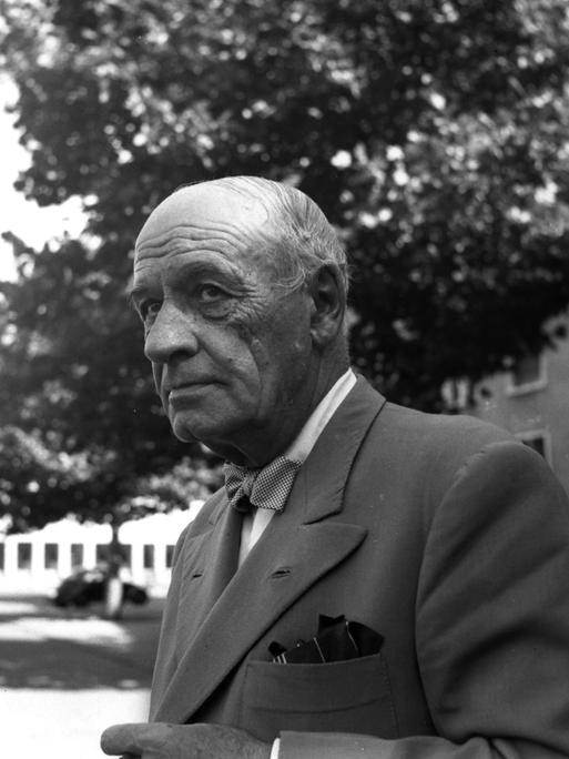 José Ortega y Gasset trägt ein graues Sakko und schaut zur Seite. Er steht vor einem Gebäude, im Hintergrund ist ein Baum und ein Auto zu sehen.