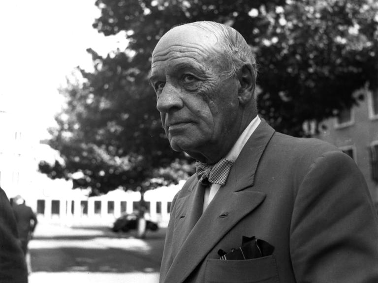 José Ortega y Gasset trägt ein graues Sakko und schaut zur Seite. Er steht vor einem Gebäude, im Hintergrund ist ein Baum und ein Auto zu sehen.