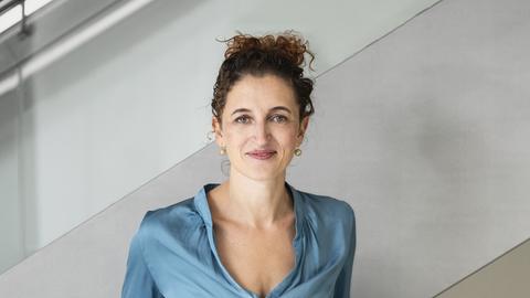 Eine junge Frau mit hochgesteckten Haaren und einer türkisblauen Bluse steht vor einem Treppenaufgang