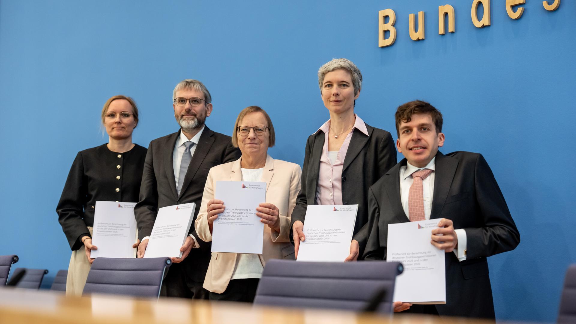 Die fünf Wisenschaftler stehen nebeneinander vor der blauen Wand der Bundespressekonferenz und halten lächelnd Exemplare ihres Berichts vor sich. 