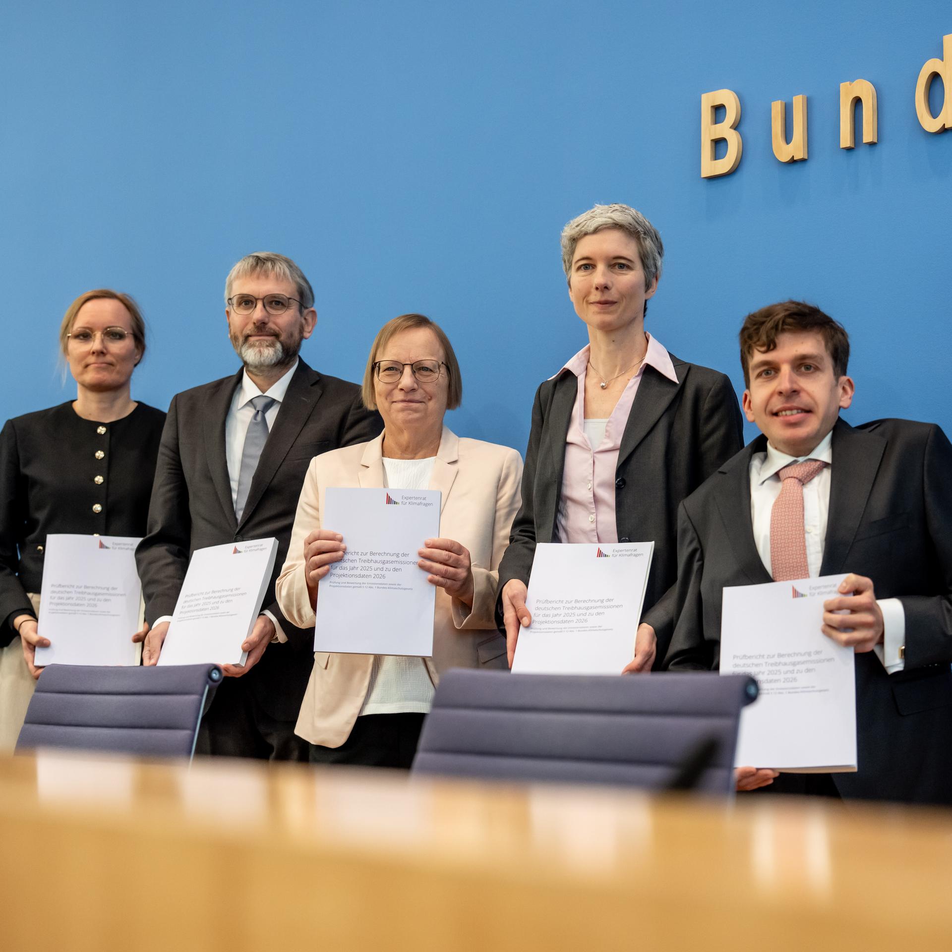 Die fünf Wisenschaftler stehen nebeneinander vor der blauen Wand der Bundespressekonferenz und halten lächelnd Exemplare ihres Berichts vor sich. 