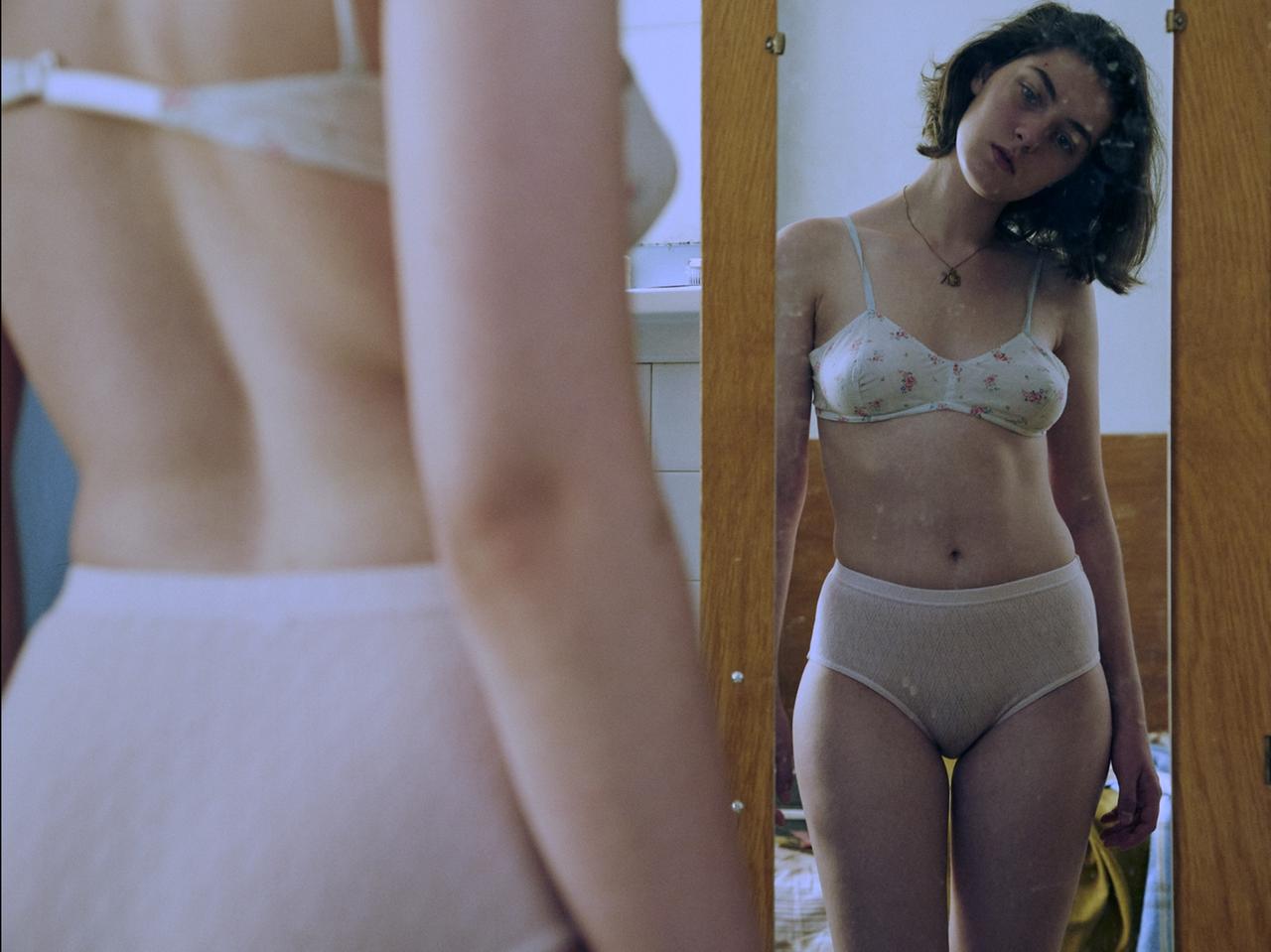 Anamaria Vartolome betrachtet sich in der Rolle der ungewollt schwangeren Anne im Spiegel. Sie trägt einen weißen BH und einen weißen Slip.