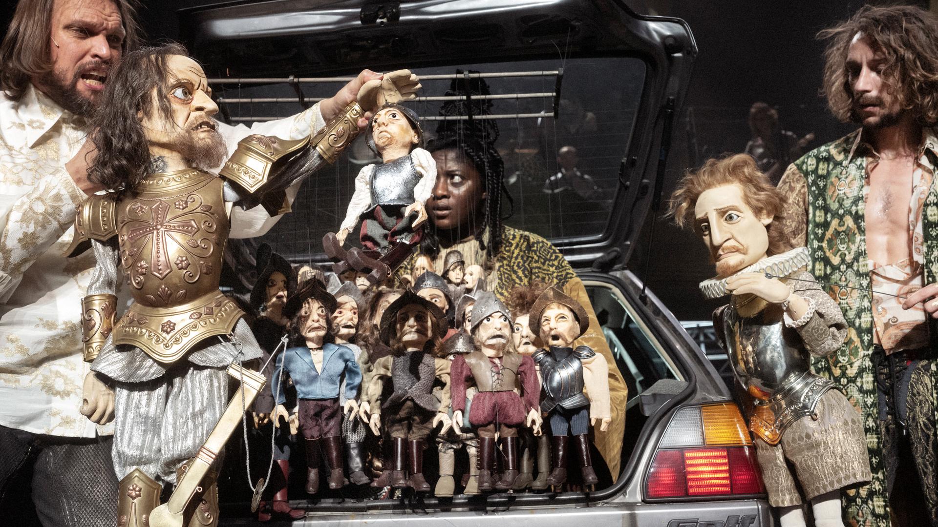 Zwei Männer bewegen Handpuppen, die aussehen wie Konquistadoren, im Gepäckraum eines VW Golfs. Eine schwarze beobachtet sie aus dem Inneren des Autos. Zwei Männer bewegen Handpuppen, die aussehen wie Konquistadoren, im Gepäckraum eines VW Golfs. Eine schwarze beobachtet sie aus dem Inneren des Autos.