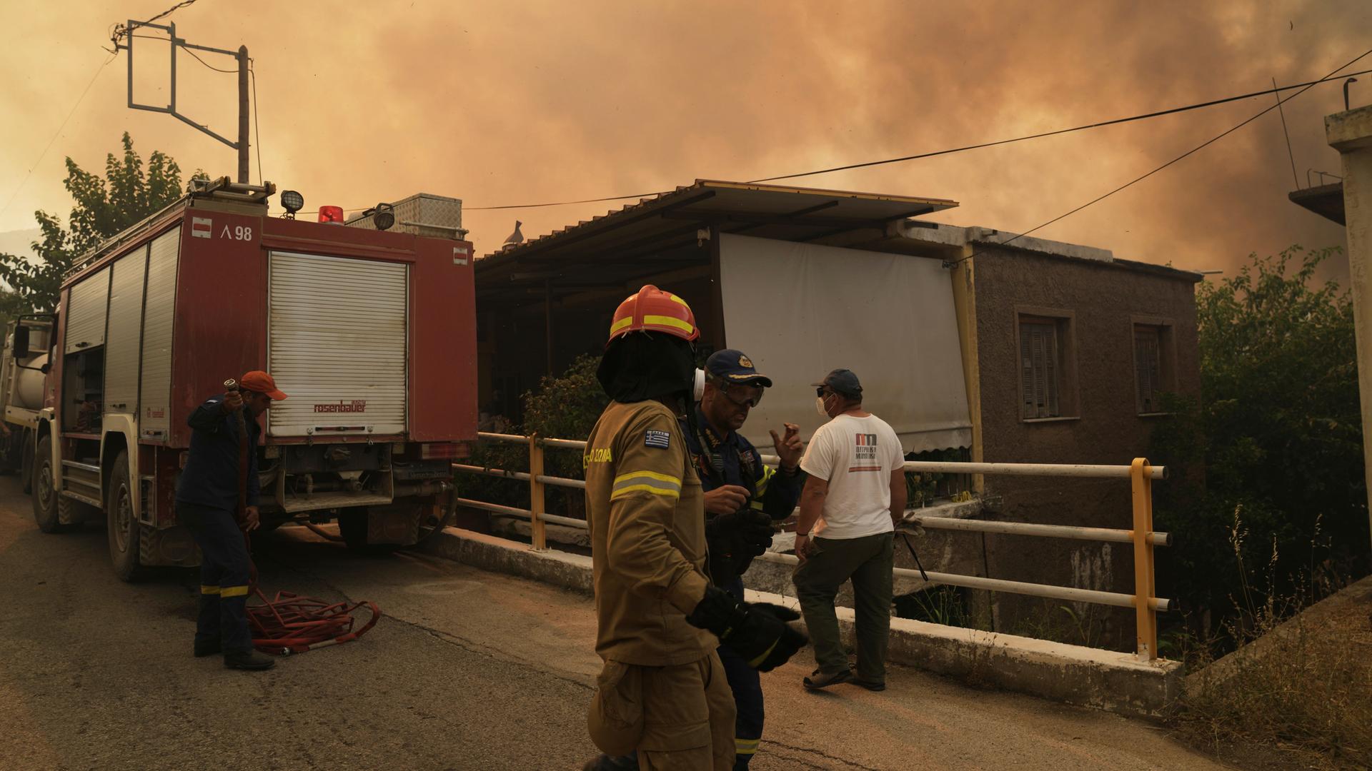 An einer Straße vor einem Haus steht ein Fahrzeug der Feuerwehr. Ein Feuerwehrmann spricht mit zwei Anwohnern. Der Himmel ist bräunlich und verraucht. An einer Straße vor einem Haus steht ein Fahrzeug der Feuerwehr. Ein Feuerwehrmann spricht mit zwei Anwohnern. Der Himmel ist bräunlich und verraucht.