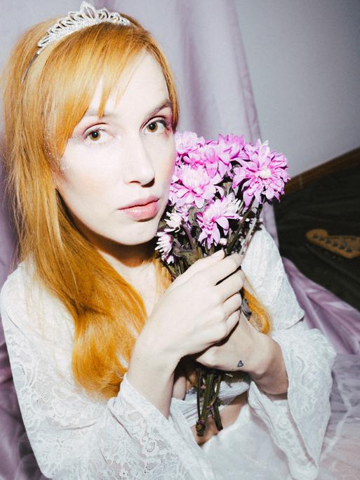 Eine junge Frau mit glatten, rotblonden Haaren sitzt auf einem Stuhl mit lila farbenem Kissen, kleiner Prinzessinnenkrone und einem Blumenstrauß in einem Raum, in dem eine Gitarre auf dem Fußboden liegt. 