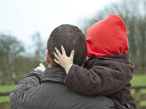 Ein Vater trägt sein Kind auf dem Arm, das Kind legt seine Hand umarmend auf des Kopf des Vaters.