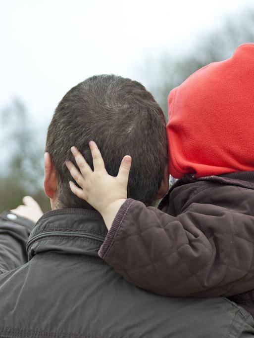 Ein Vater trägt sein Kind auf dem Arm, das Kind legt seine Hand umarmend auf des Kopf des Vaters.