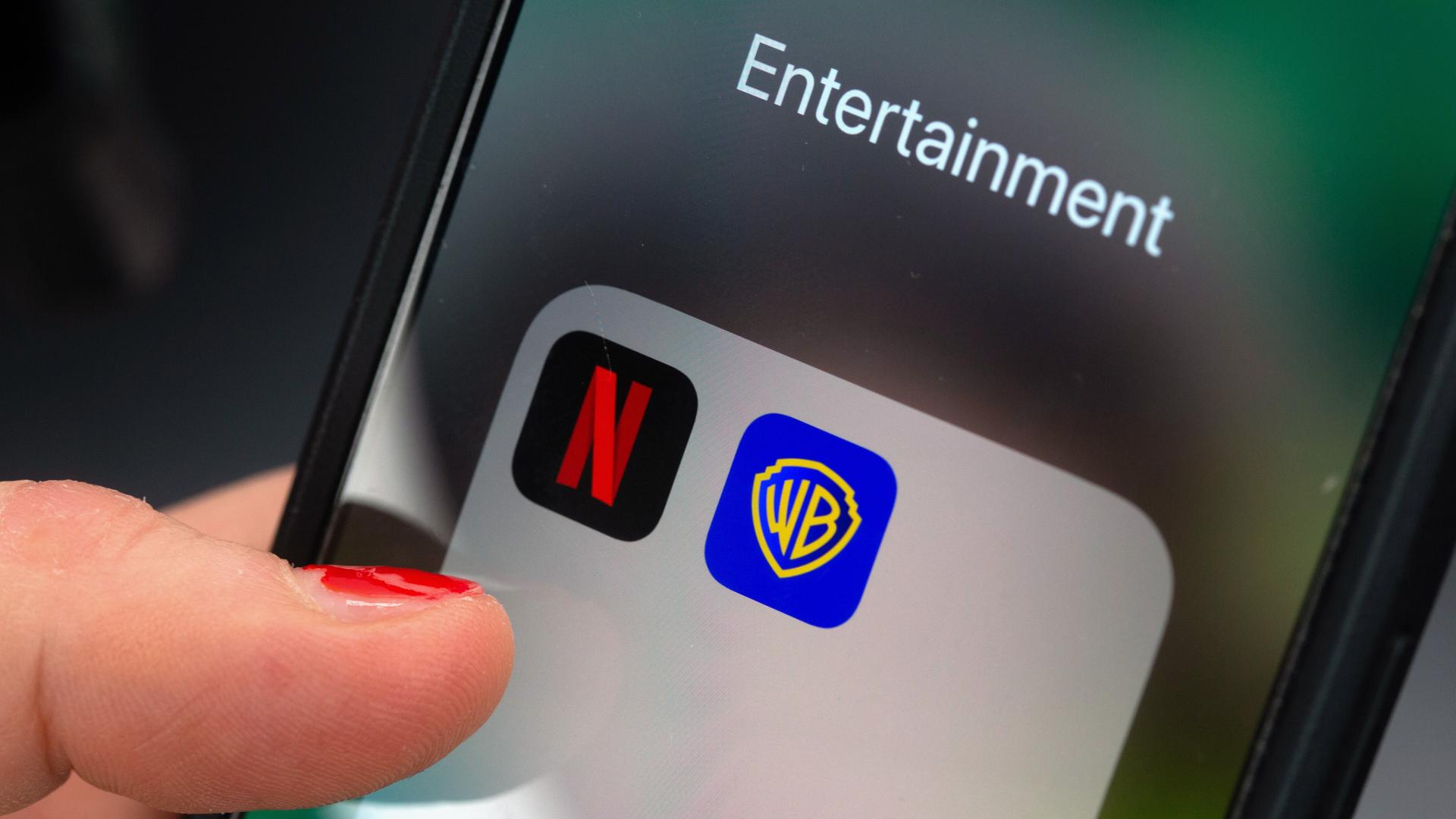 Ein Finger tippt auf einem Smartphone auf ein App-Ordnerfeld mit den Logos der Streamingplattform Netflix (links) und des Filmstudios Warner Brothers (rechts). 