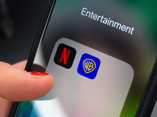 Ein Finger tippt auf einem Smartphone auf ein App-Ordnerfeld mit den Logos der Streamingplattform Netflix (links) und des Filmstudios Warner Brothers (rechts). 