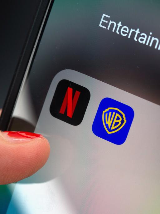 Ein Finger tippt auf einem Smartphone auf ein App-Ordnerfeld mit den Logos der Streamingplattform Netflix (links) und des Filmstudios Warner Brothers (rechts). 