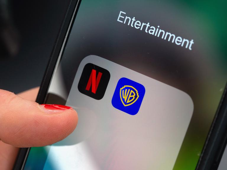Ein Finger tippt auf einem Smartphone auf ein App-Ordnerfeld mit den Logos der Streamingplattform Netflix (links) und des Filmstudios Warner Brothers (rechts). 