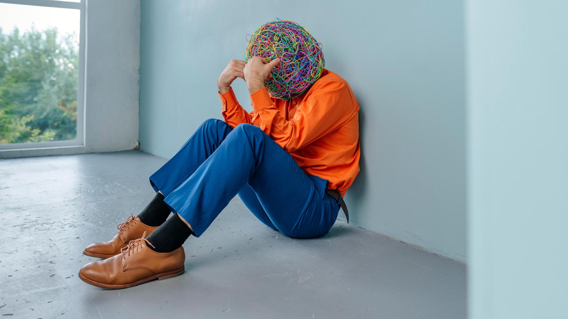 Lonely businessman with mask of chaotic thoughts sitting with head in hands near blue wall || Modellfreigabe vorhanden