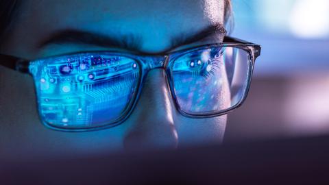 Eine Frau mit Brille sitzt vor einem Computer. In ihren Brillengläsern spiegelt sich bläulich der Aufbau eines Chips.