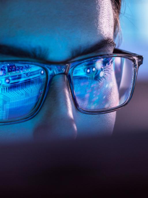 Eine Frau mit Brille sitzt vor einem Computer. In ihren Brillengläsern spiegelt sich bläulich der Aufbau eines Chips.