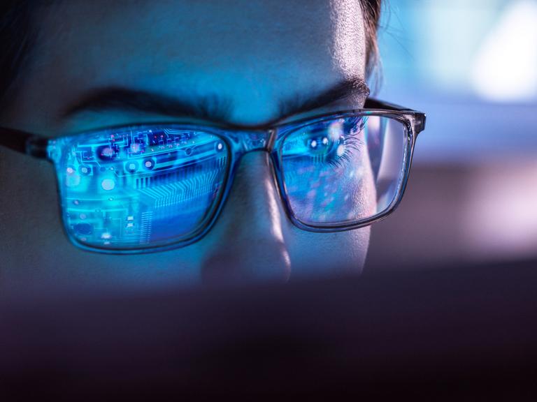Eine Frau mit Brille sitzt vor einem Computer. In ihren Brillengläsern spiegelt sich bläulich der Aufbau eines Chips.