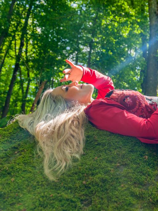 Anke Helfrich liegt mit offenen blonden Haaren mitten im Wald auf einer Böschung, während ein Sonnenstrahl direkt ihr Gesicht trifft. 