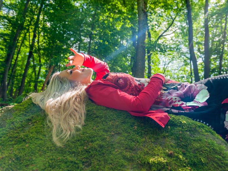 Anke Helfrich liegt mit offenen blonden Haaren mitten im Wald auf einer Böschung, während ein Sonnenstrahl direkt ihr Gesicht trifft. 