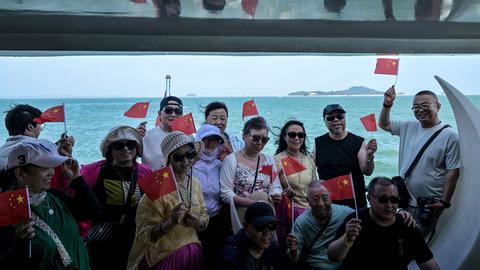 Chinesische Touristen posieren auf einem Boot mit kleinen chinesischen Flaggen. Im Hintergrund sind die Umrisse der Kinmen-Inseln zu sehen,