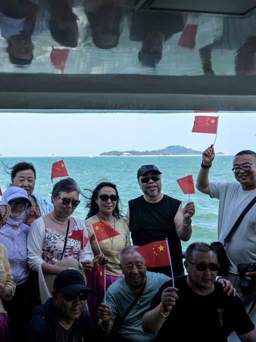 Chinesische Touristen posieren auf einem Boot mit kleinen chinesischen Flaggen. Im Hintergrund sind die Umrisse der Kinmen-Inseln zu sehen, Chinesische Touristen posieren auf einem Boot mit kleinen chinesischen Flaggen. Im Hintergrund sind die Umrisse der Kinmen-Inseln zu sehen,