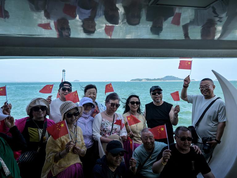 Chinesische Touristen posieren auf einem Boot mit kleinen chinesischen Flaggen. Im Hintergrund sind die Umrisse der Kinmen-Inseln zu sehen, Chinesische Touristen posieren auf einem Boot mit kleinen chinesischen Flaggen. Im Hintergrund sind die Umrisse der Kinmen-Inseln zu sehen,