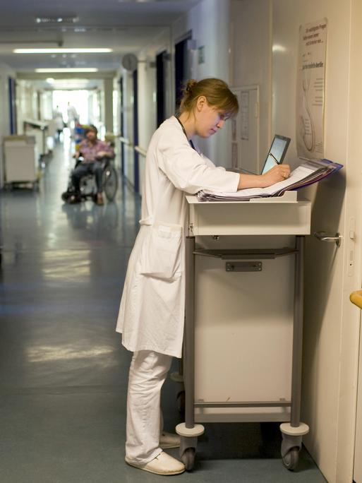 Blick in einen Krankenhausflur, in dem eine Ärztin etwas in ihren Unterlagen notiert. 