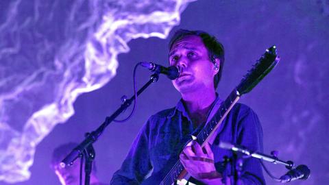 Daniel Rossen steht auf der Bühne am Mikro und spielt Gitarre. Die Beleuchtung taucht den Raum in lila Licht.