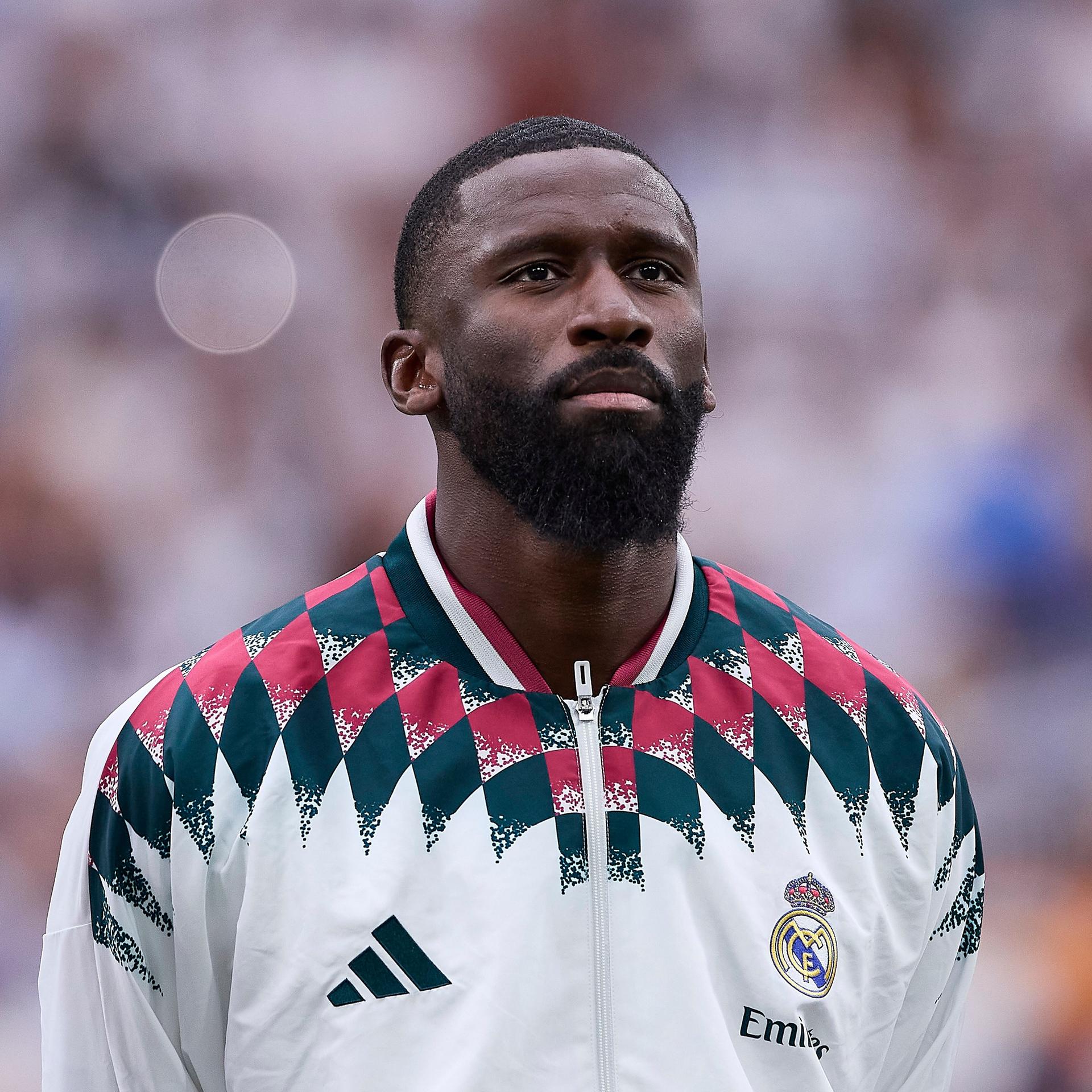 Antonio Rüdiger steht zwischen zwei anderen Fußballspielern, er trägt eine weiße Trikotjacke.