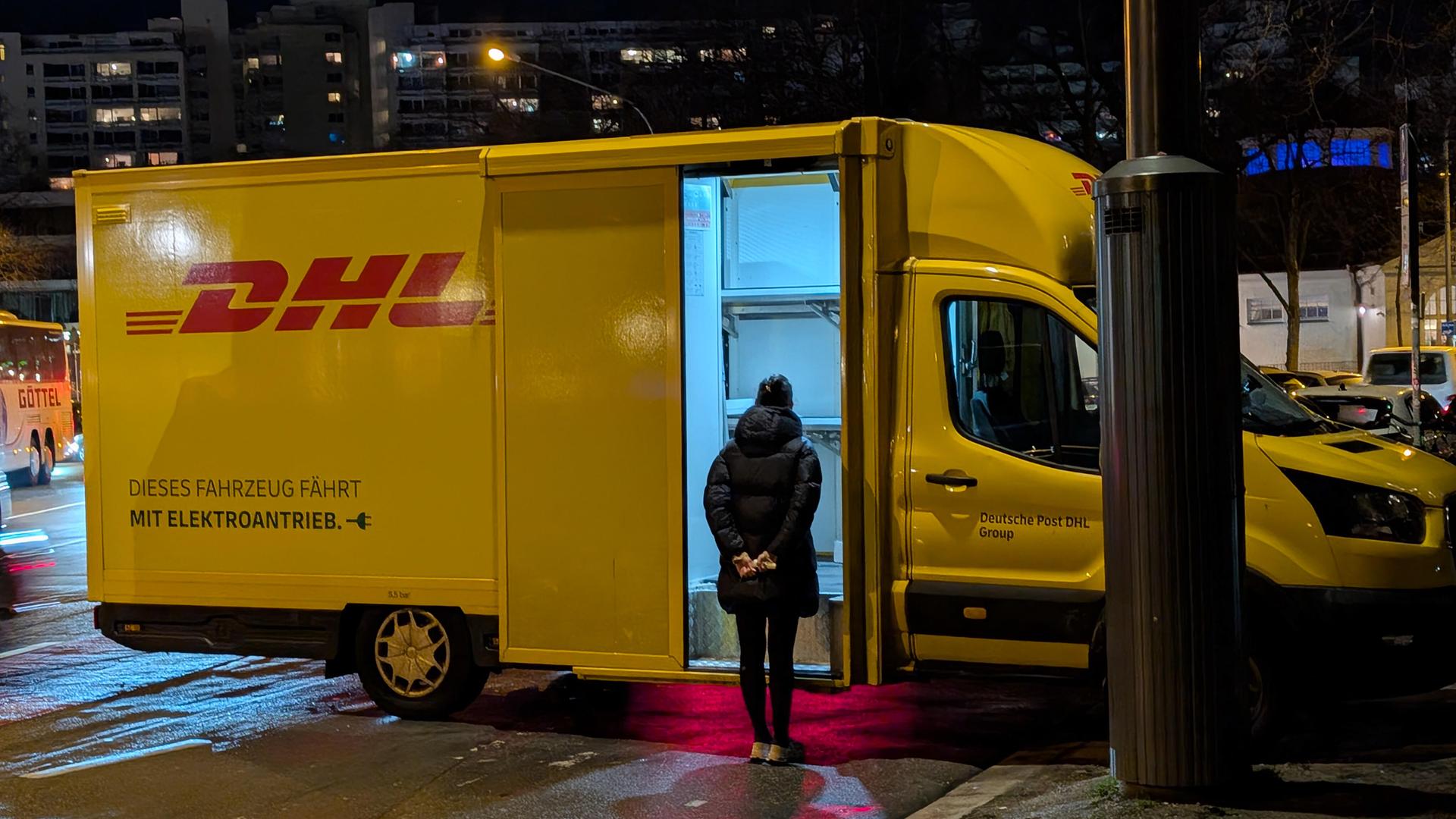 Eine Frau steht vor einer geöffneten Ladetür eines gelben DHL-Transporters, im Hintergrund nächtliche Stadtkulisse.