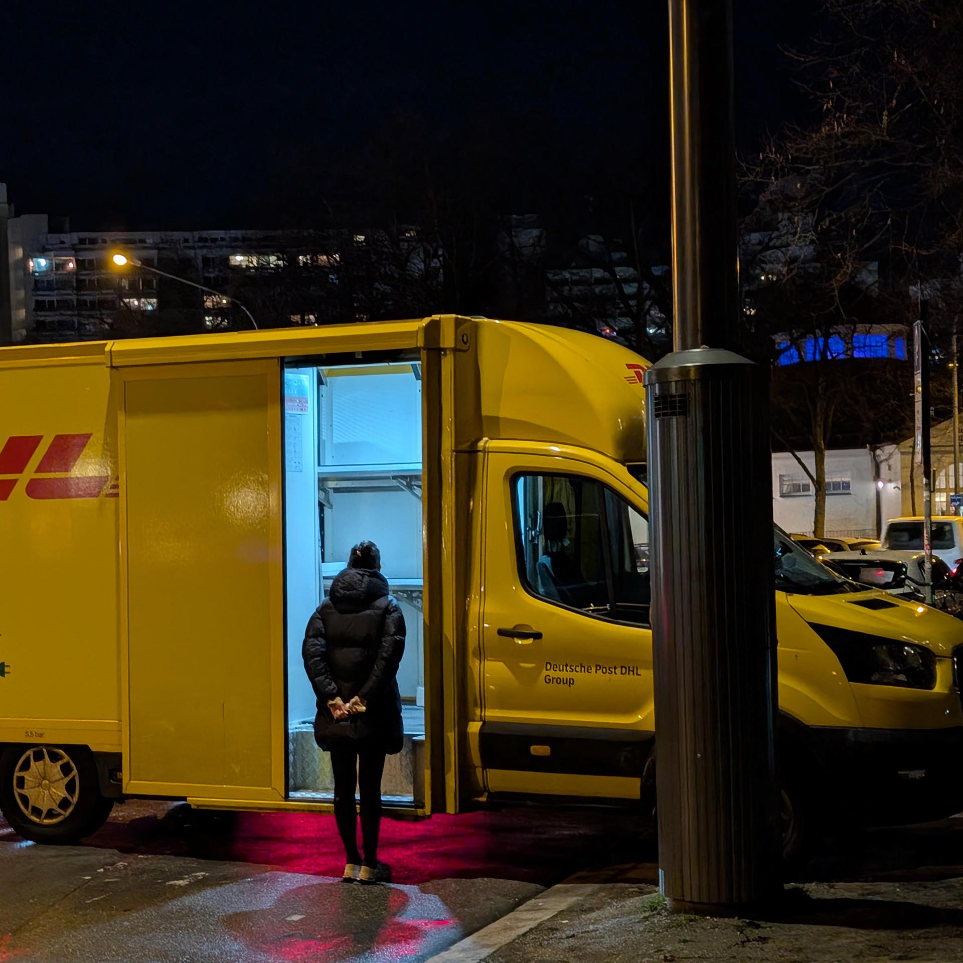 Eine Frau steht vor einer geöffneten Ladetür eines gelben DHL-Transporters, im Hintergrund nächtliche Stadtkulisse.