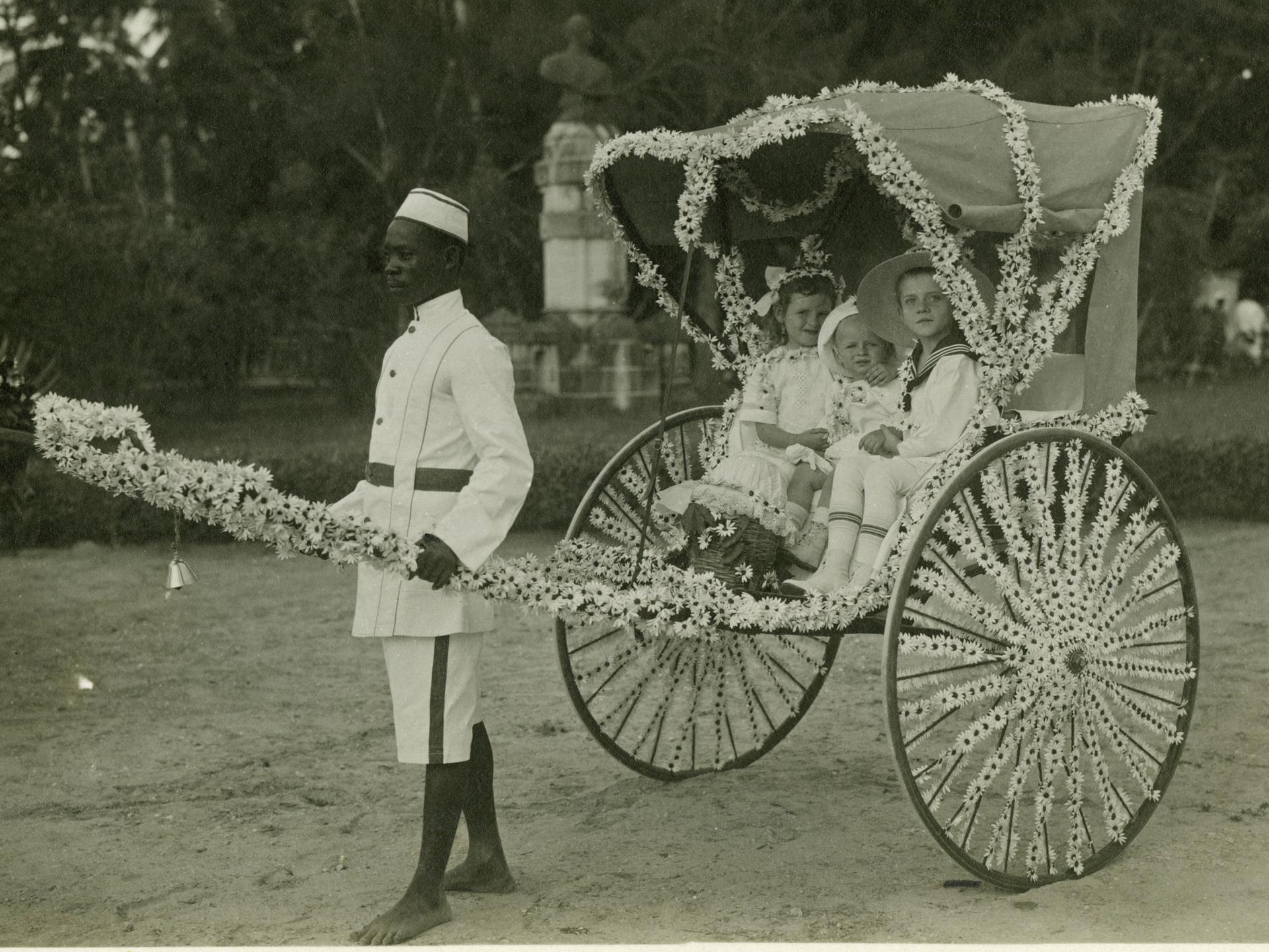 Ein Diener zieht eine mit Blumen geschmückte Rikscha mit drei weißen Kindern am vom Frauenverein des Roten Kreuzes organisierten "Margaritentag" in Daressalam (1912).