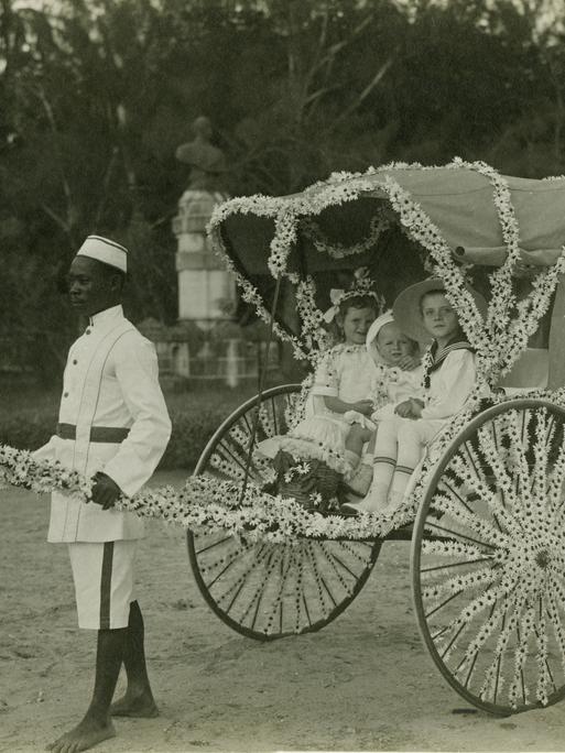 Ein Diener zieht eine mit Blumen geschmückte Rikscha mit drei weißen Kindern am vom Frauenverein des Roten Kreuzes organisierten "Margaritentag" in Daressalam (1912).