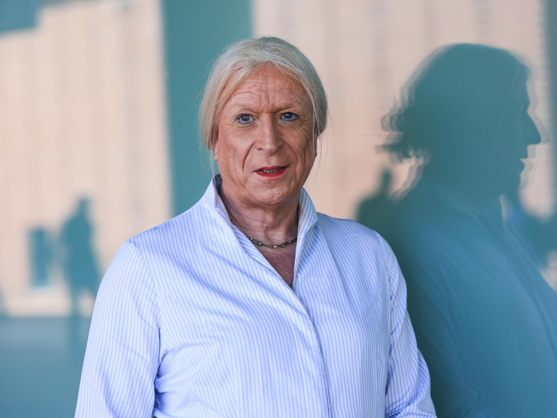 Georgine Kellermann trägt eine hellblau gestreifte Bluse die langen weißblonden Haare zu einem Zopf. Hinter sich wirft sie einen Schatten im Profil an die Wand.