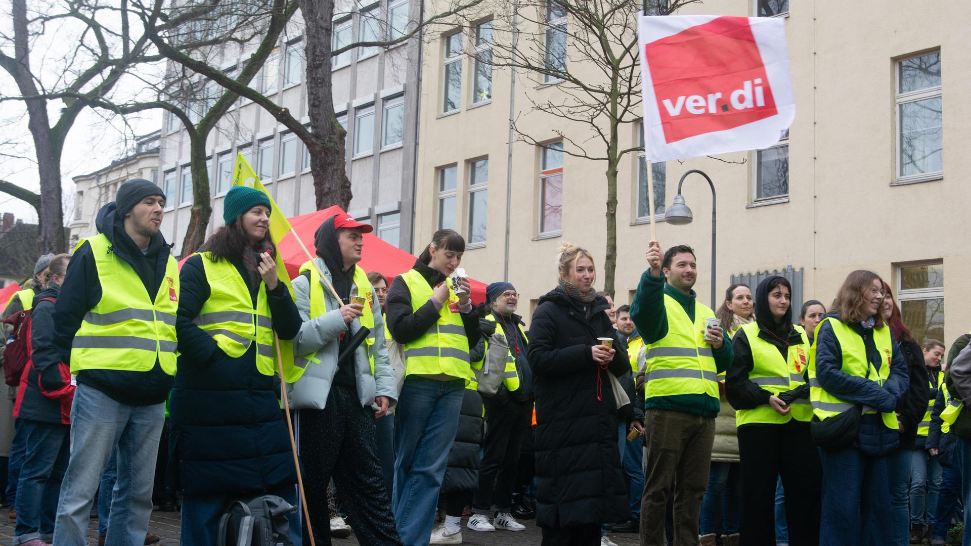 Eine Gruppe Streikender - viele davon in gelben Warnwesten - stehen auf einem Platz mit Bäumen. Im Hintergrund Wohnhäuser. 