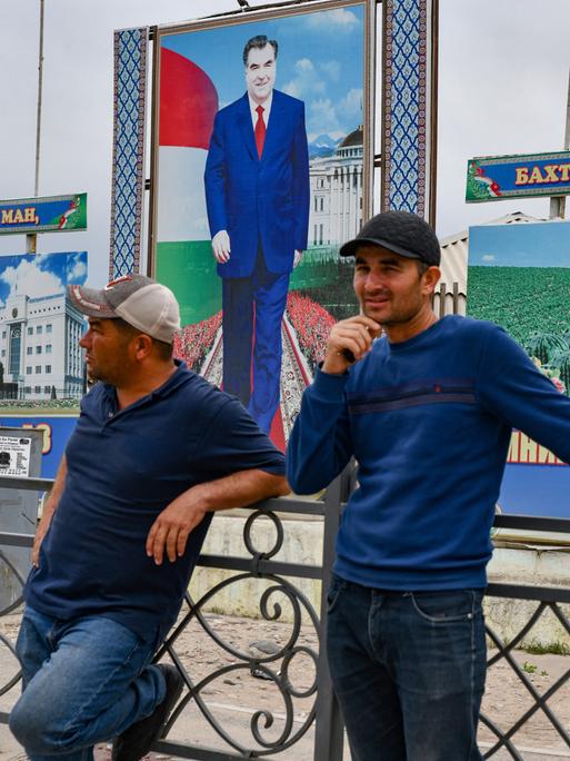 Zwei Männer stehen plaudernd auf der straße vor einem stilisierten Porträt des tadschikischen Präsidenten Emomali Rahmon.