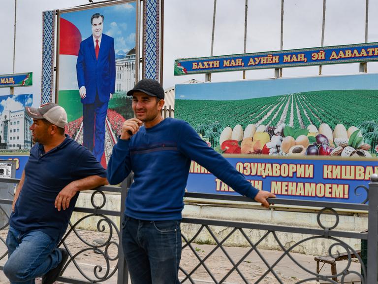 Zwei Männer stehen plaudernd auf der straße vor einem stilisierten Porträt des tadschikischen Präsidenten Emomali Rahmon.