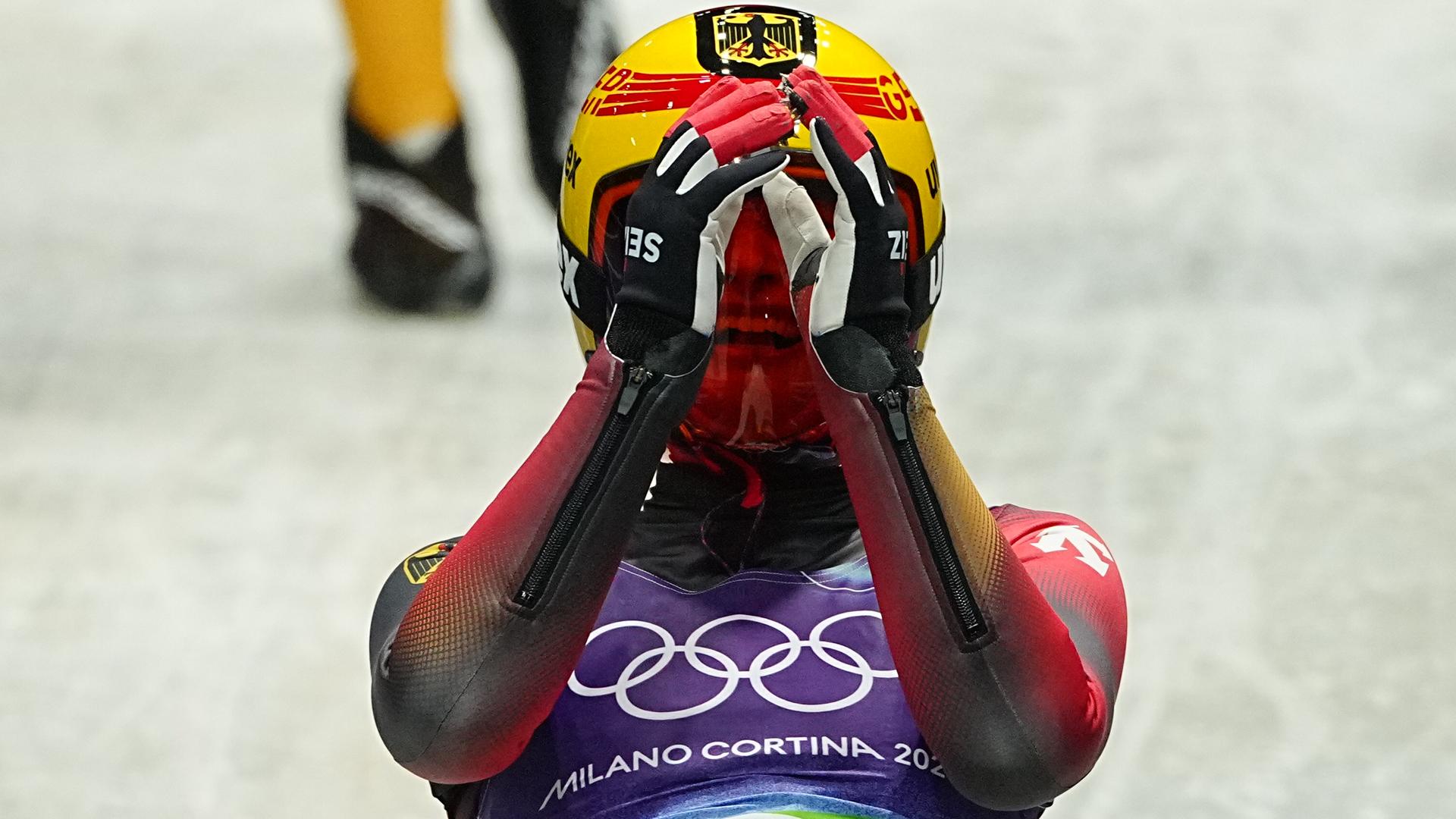 Taubitz sitzt auf dem Schlitten und hält beide Hände vorm das Visier ihres Helms. Dahinter unscharf die Eisbahn und darauf die Füße einer Person.