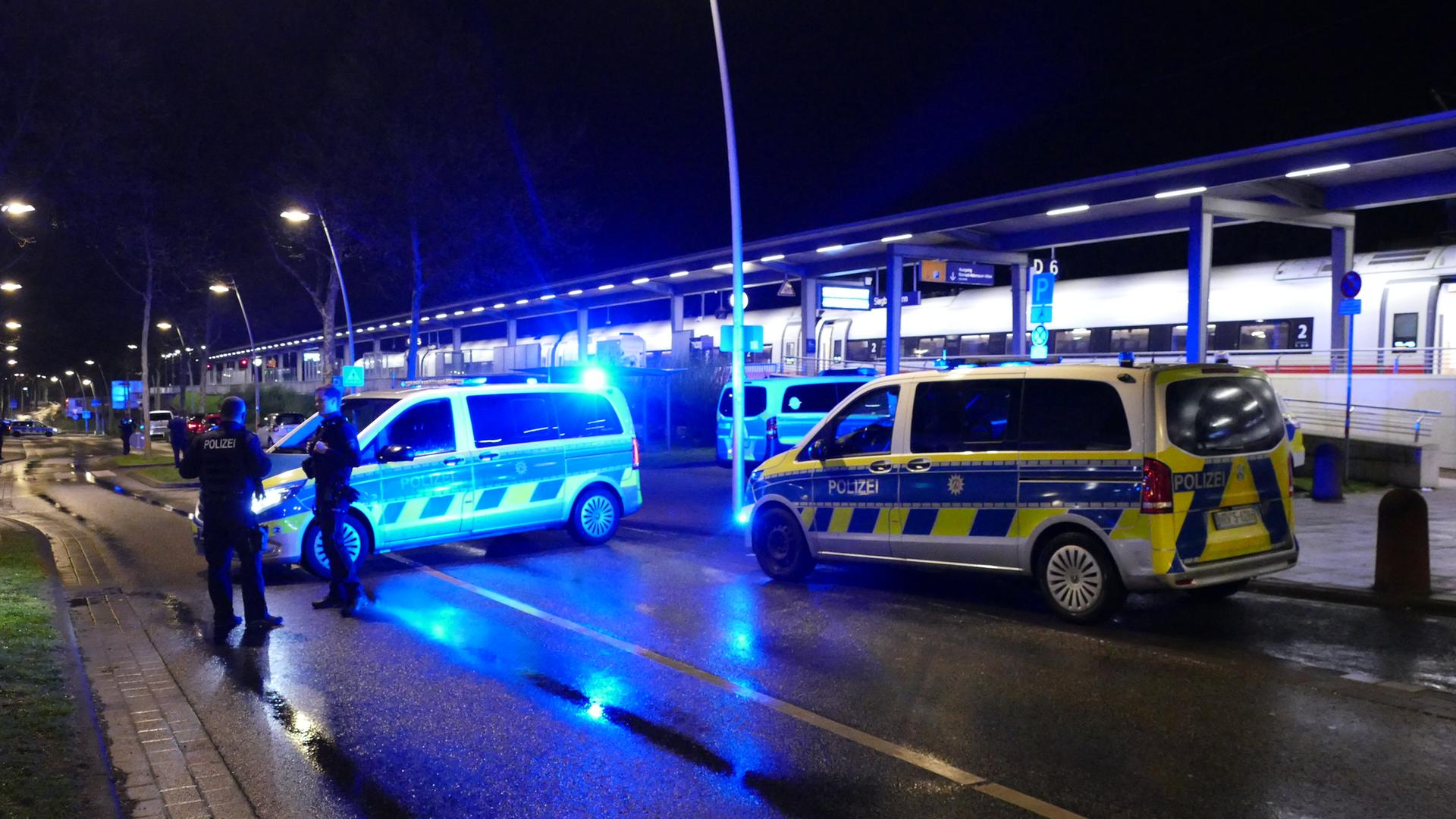 Zwei Polizeiwagen und zwei Polizisten stehen in der Dunkelheit auf einer Straße. Dahinter ist ein ICE an einer Station zu sehen. Zwei Polizeiwagen und zwei Polizisten stehen in der Dunkelheit auf einer Straße. Dahinter ist ein ICE an einer Station zu sehen.