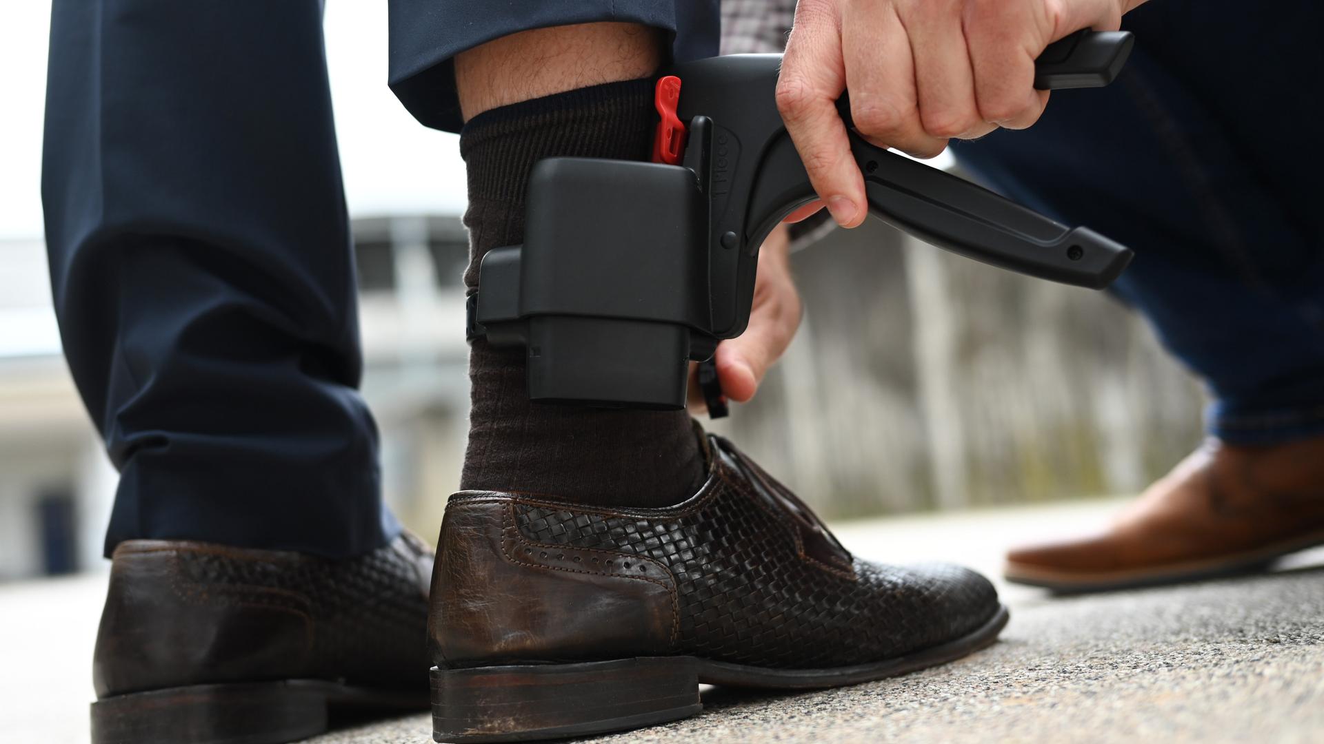 Ein Mann entfernt eine elektronische Fußfessel - einen kleinen schwarzen Kasten, etwa so groß wie ein Handy - an dem Bein eines anderen Mannes.