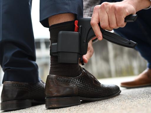 Ein Mann entfernt eine elektronische Fußfessel - einen kleinen schwarzen Kasten, etwa so groß wie ein Handy - an dem Bein eines anderen Mannes.