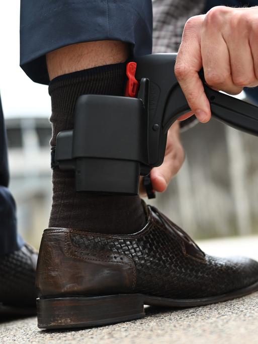 Ein Mann entfernt eine elektronische Fußfessel - einen kleinen schwarzen Kasten, etwa so groß wie ein Handy - an dem Bein eines anderen Mannes.