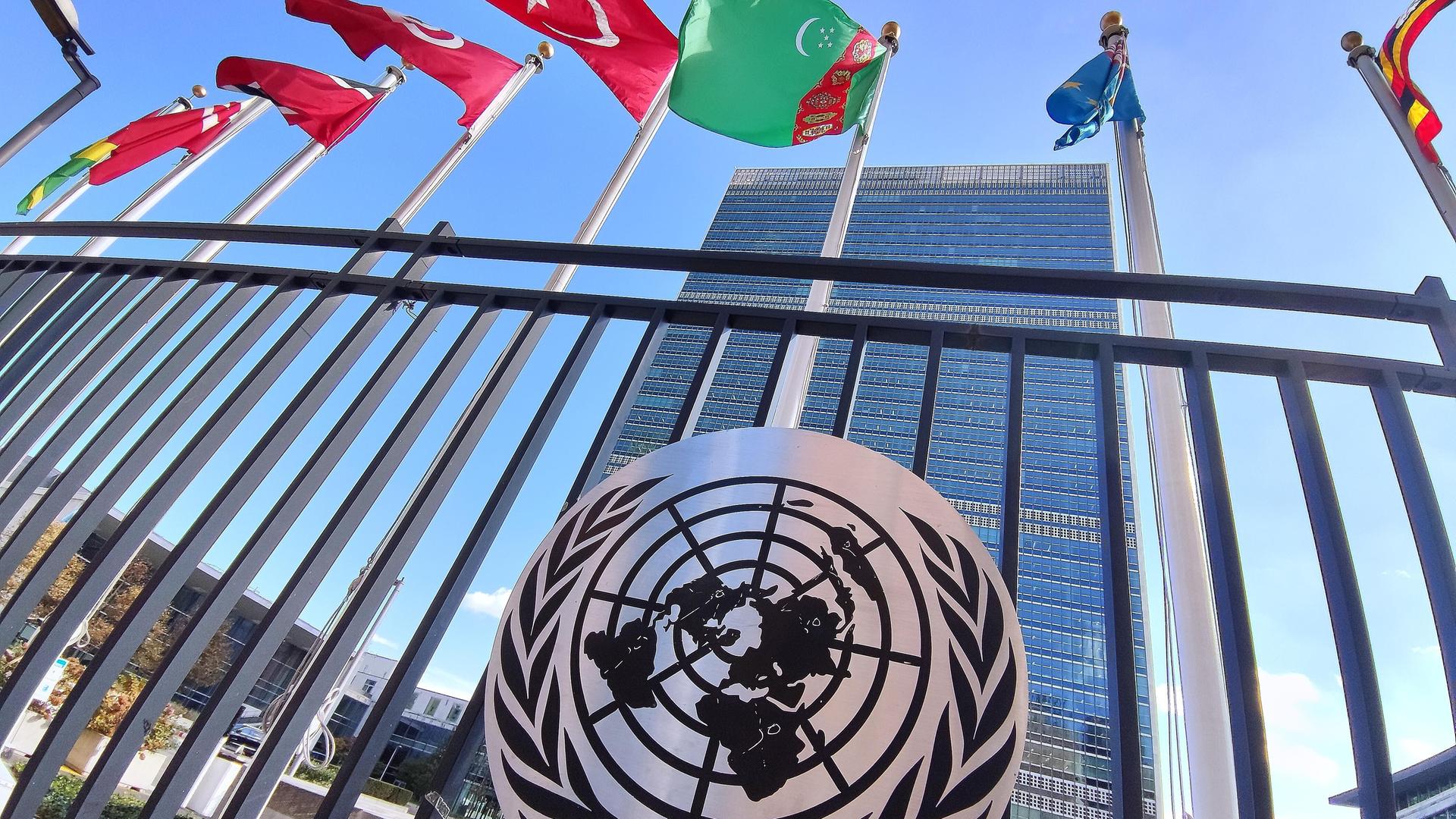 Blick auf das Gebäude der Vereinten Nationen in New York. Am Zaun ist der Logo der UN angebracht, darüber wehen die Flaggen diverser Staaten.
