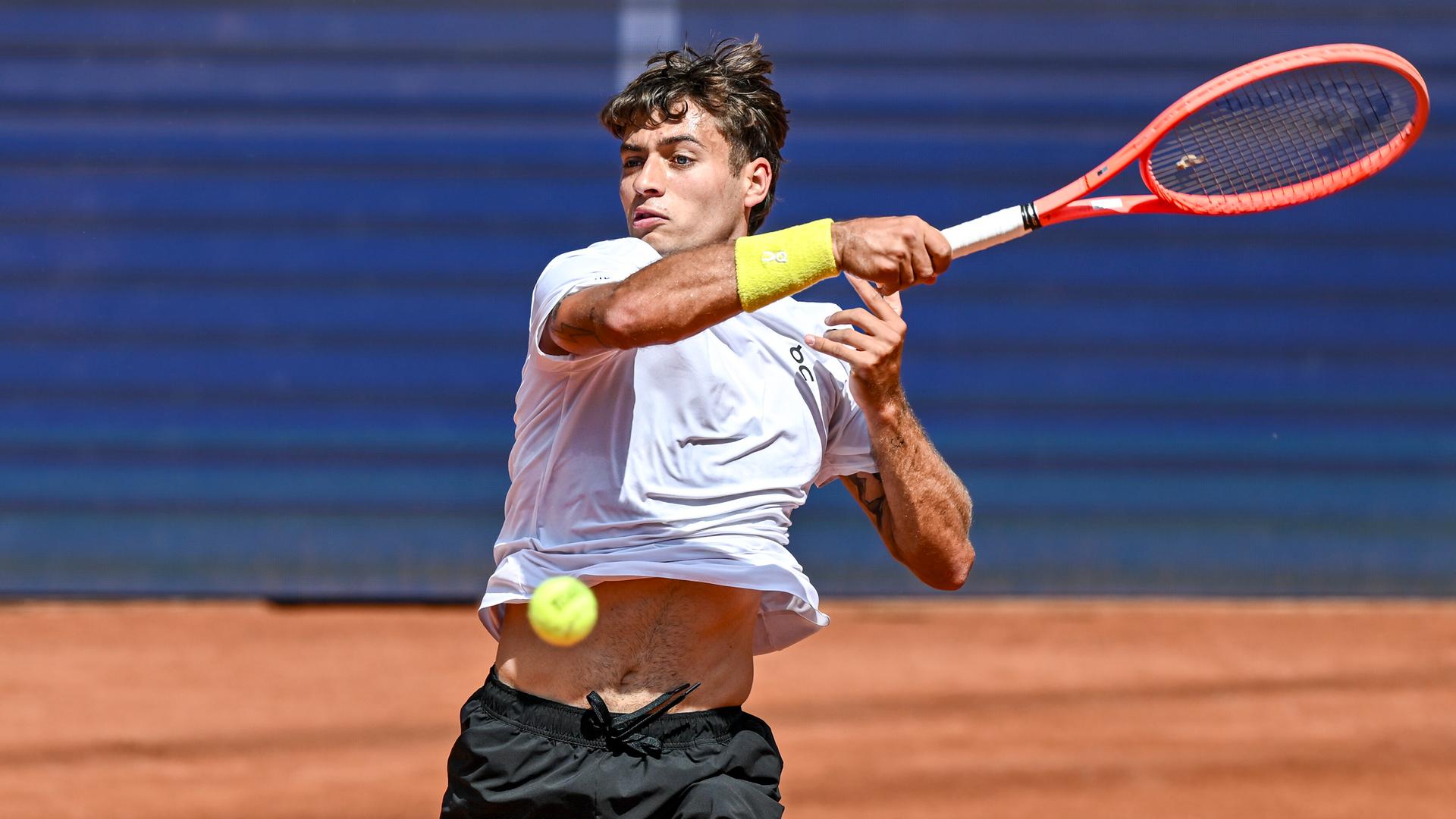 Cobolli holt mit einem roten Tennisschläger zum Schlag aus. Er trägt ein weißes Trikot und eine schwarze Hose. Cobolli holt mit einem roten Tennisschläger zum Schlag aus. Er trägt ein weißes Trikot und eine schwarze Hose.